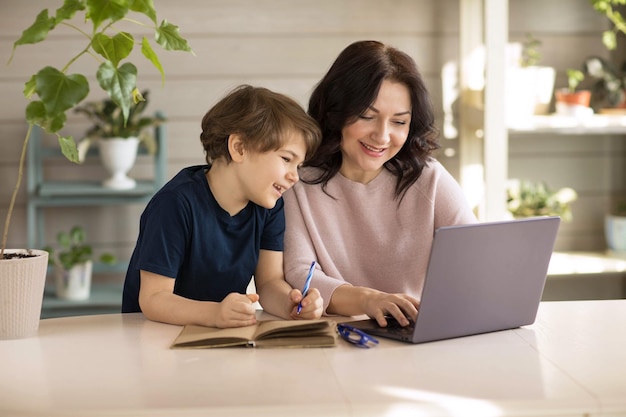 Moeder en zoon zijn bezig met online school, zitten op een laptop en maken aantekeningen in een notitieboekje
