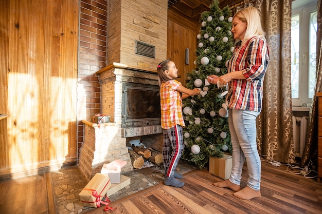 Moeder houdt een dochtertje bij de kerstboom op een feestelijke avond in een rustiek houten huis, Nieuwjaar, wintertijd