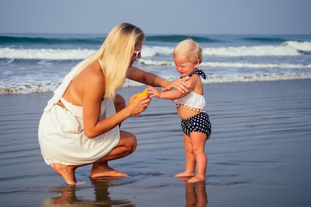 Moeder in stijlvolle bril en witte jurk zet oranje spuitfles SPF op pasgeboren baby van een jaar oude dochter een zonnebrandcrème op het strand