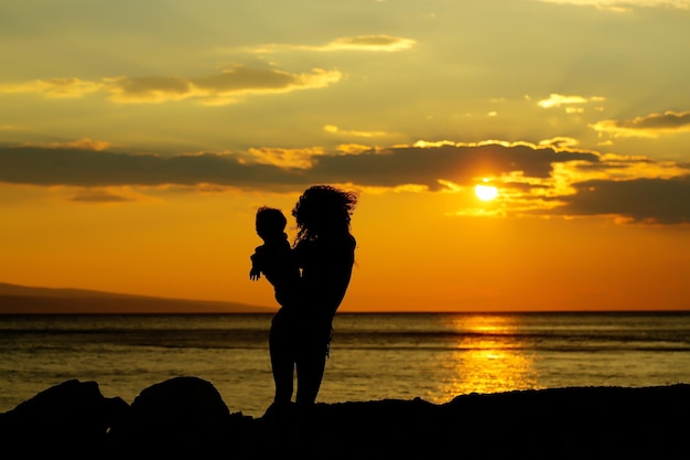 Moeder jonge vrouw en zoon babyjongen silhouetten speelt op strand in de buurt van zee of oceaanwater in avond of schemering zonsondergang zomerdag op natuurlijke achtergrond