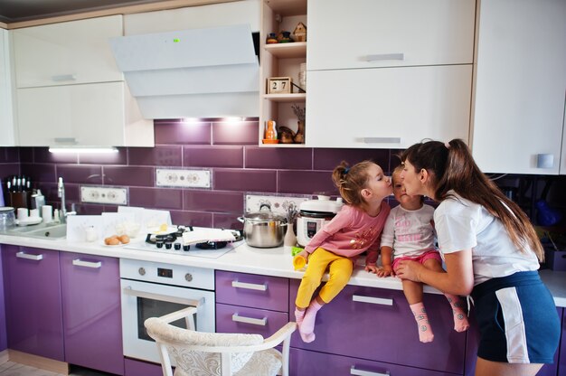 Moeder kust haar meisjeskinderen in de keuken, gelukkige kindermomenten. Moeder en twee dochters.