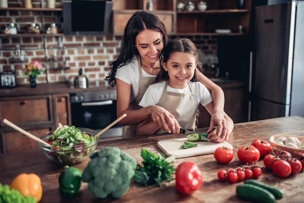 Moeder leert haar dochter komkommer te snijden voor de salade in de keuken en kijkt naar de camera