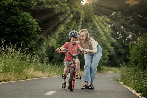 Moeder leert zoon fietsen Gelukkige schattige jongen in helm leert fietsen op het fietspad in de zomer regenachtige dag bij zonsondergang Familieweekend