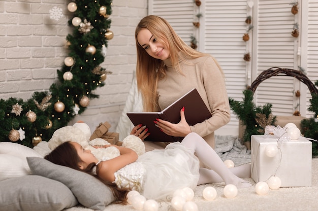 Moeder leesboek aan haar dochtertje in de buurt van de kerstboom thuis