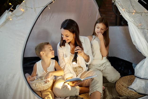 Moeder leest een boek met sprookjes voor haar kinderen terwijl ze 's nachts in een tent zit. Moeder zoon en dochter lezen een boek met een zaklamp in hun handen
