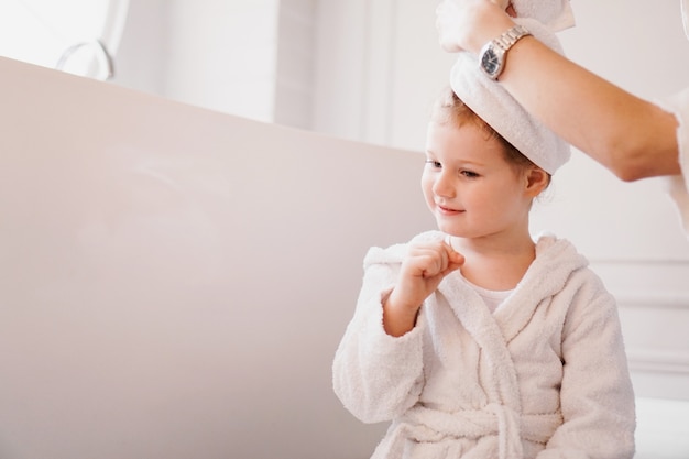 Moeder legt een handdoek op het haar van haar dochter. Dochtertje in een witte badjas in een lichte badkamer.