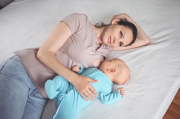 Moeder ligt met pasgeboren baby baby in blauwe jumpsuit knuffelen en geeft hem borstvoeding met moedermelk