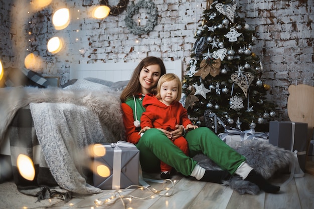 Moeder met dochter in rode warme truien springen op het bed. Gelukkig moederschap. Warme familierelaties. Kerst- en nieuwjaarsinterieur. Dol zijn op. Familieconcept.