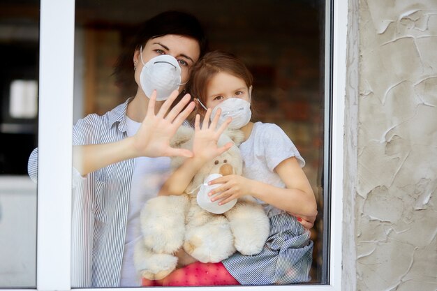 Moeder met dochterkind in masker die van venster, coronavirus kijken.