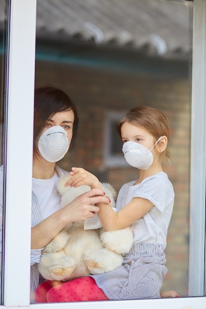 Moeder met dochterkind in masker die van venster, coronavirus kijken.