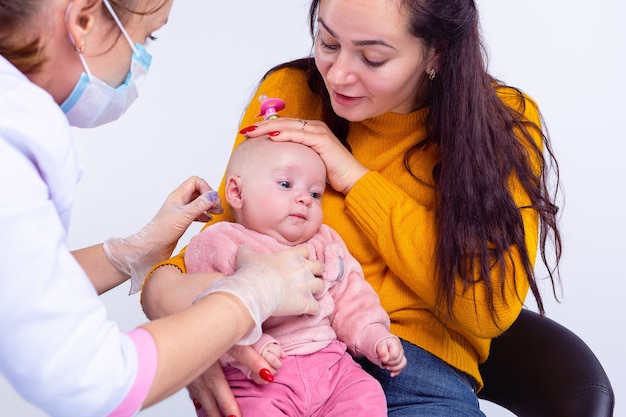 Moeder met een babymeisje zittend op een stoel dokter vrouw onderzoekt de baby in een roos die jurk draagt moeder houdt een baby op haar knieën