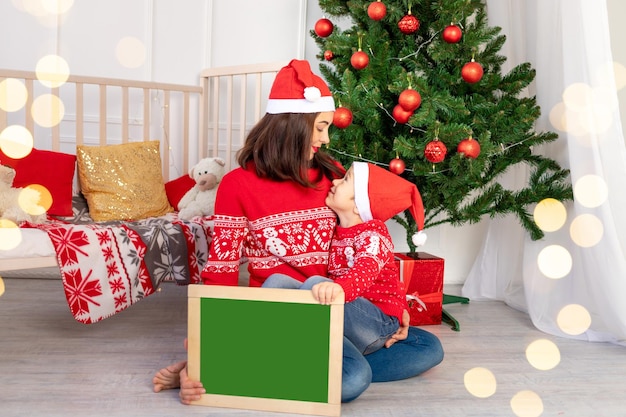 Moeder met een kind in rode truien in de kinderkamer bij de kerstboom met een nieuwjaarsinterieur houdt een bord vast met ruimte voor tekst