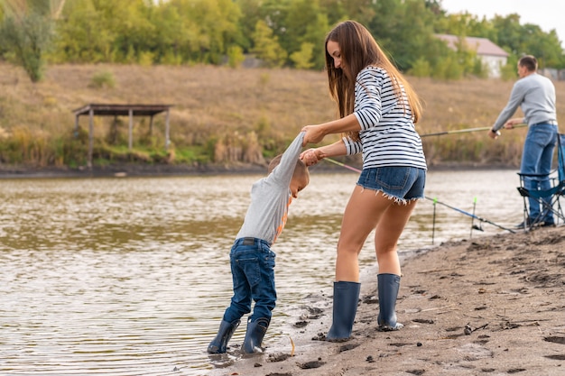 Moeder met een kleine zoon loopt in rubberen laarzen langs de zandige oever van het meer. Rondhangen met kinderen in de natuur, weg van de stad