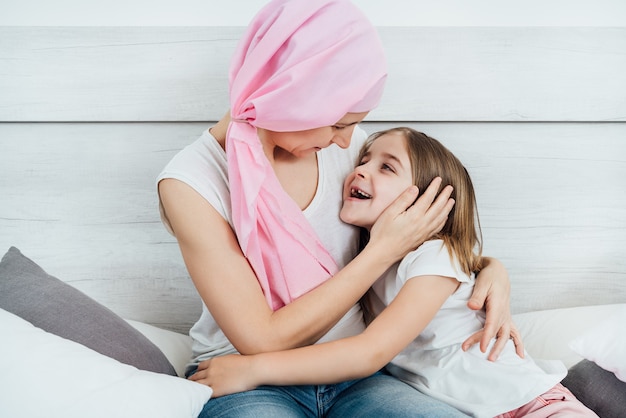 Moeder met kanker draagt een roze hoofddoek die het gezicht van haar dochter knuffelt en streelt met prachtig blond haar. Ze zitten allebei vrolijk in bed tegen een witte achtergrond