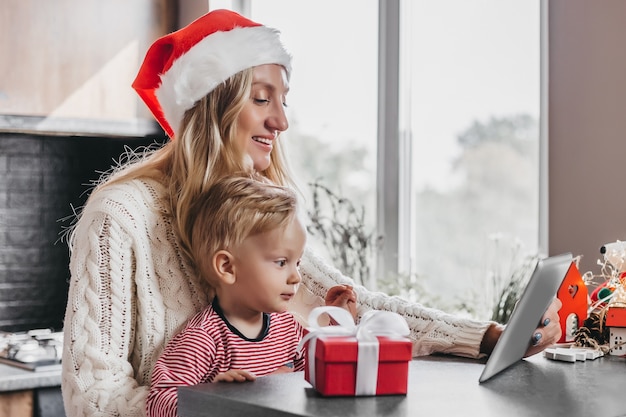 Moeder met kerstmuts en zoontje zitten aan tafel en houden een tablet in hun handen en praten via videoconferentie