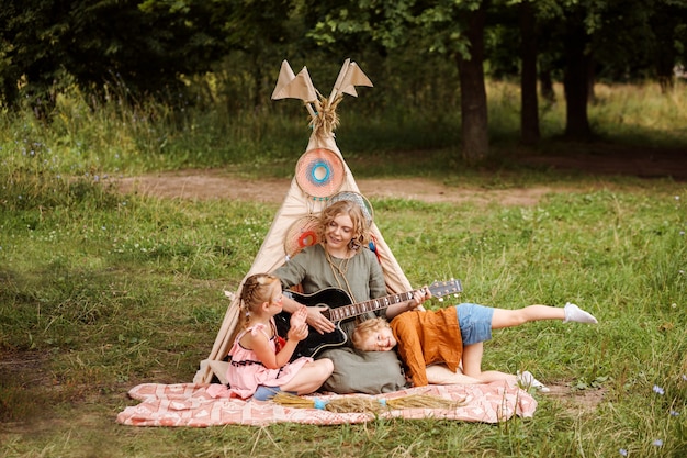 Moeder speelt gitaar voor haar kleine dochters naast wigwam decoratie buiten in de zomer. Samen de zomervakantie doorbrengen.