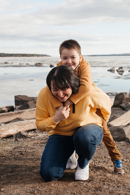 Moeder speelt met haar zoon op de rivieroever. Een mooie moeder en zoon in gele hoodies hebben plezier met spelen en lachen op een zonnige dag. Buitenrecreatie.