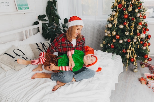 Moeder speelt met haar zoon op het bed thuis bij de kerstboom. vrouw en kind in santa claus hoeden. Kerstmis.