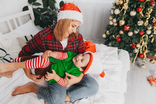 Moeder speelt met haar zoon op het bed thuis bij de kerstboom. vrouw en kind in santa claus hoeden. Kerstmis.