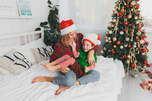 Moeder speelt met haar zoon op het bed thuis bij de kerstboom. vrouw en kind in santa claus hoeden. Kerstmis.