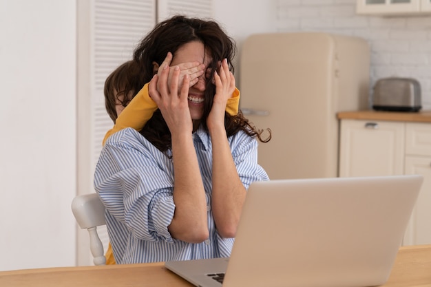 Moeder werkt thuis op laptop tijdens lockdown, kind leidt af van het werk dat de ogen van haar moeder bedekt