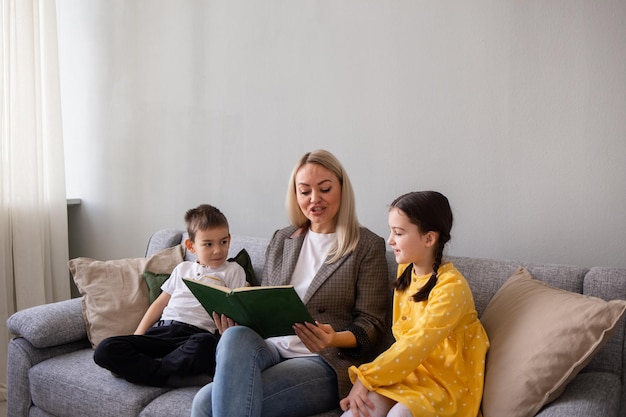 Moeder zit op een grijze bank in de kamer en leest een boek voor aan twee kinderen Thuisonderwijs