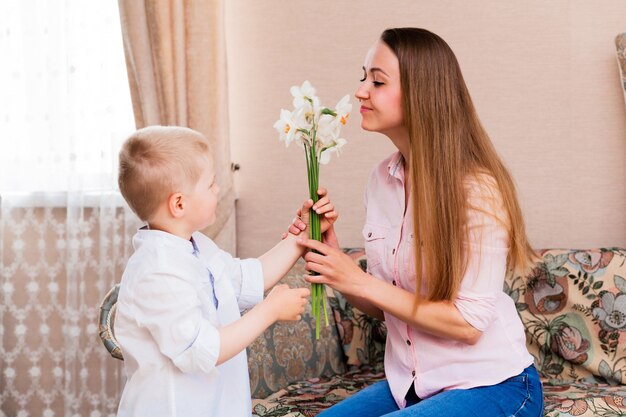 Moederdag, vakantie en familieconcept - gelukkig zoontje geeft thuis bloemen aan zijn lachende moeder. Een kind geeft mama een boeket narcissen