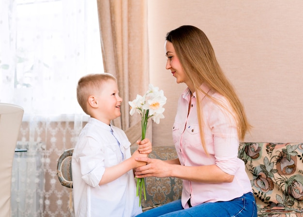 Moederdag, vakantie en familieconcept - gelukkig zoontje geeft thuis bloemen aan zijn lachende moeder. Een kind geeft mama een boeket narcissen