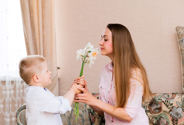 Moederdag, vakantie en familieconcept - gelukkig zoontje geeft thuis bloemen aan zijn lachende moeder. Een kind geeft mama een boeket narcissen