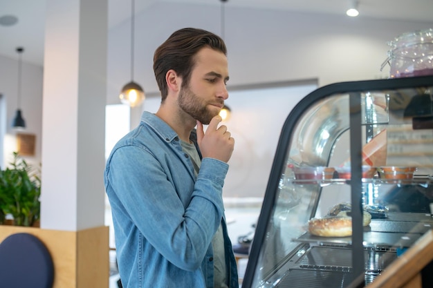 Moeilijk kiezen. Jonge, bebaarde man die zijwaarts naar de camera staat en zijn kin aanraakt, nadenkend naar voedselvertoning in café