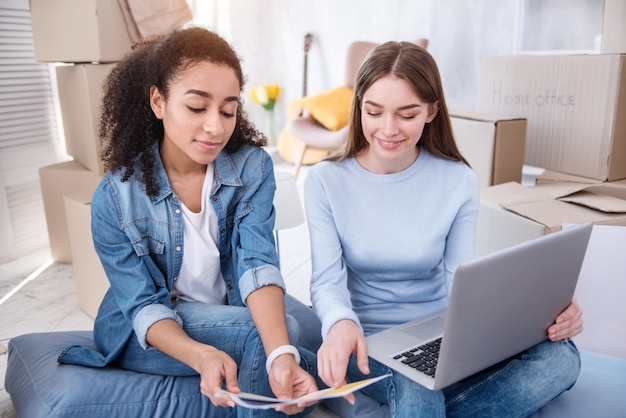 Moeilijke keuze. Aangename jonge vrouwen die in hun nieuwe appartement op de grond zitten en de kleur voor de muren uitkiezen