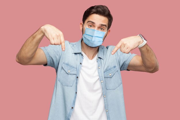 Moet hier en nu zijn. Portret van een serieuze man met een chirurgisch medisch masker in een blauw casual stijlshirt dat staat, naar beneden wijst en naar de camera kijkt. indoor studio-opname, geïsoleerd op roze achtergrond