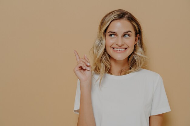 Moet je zien. Positieve jonge vrouw klant met blond haar, gekleed in witte t-shirt met aangename glimlach wijzend op kopieerruimte op oranje achtergrond. Reclame- en promotieconcept