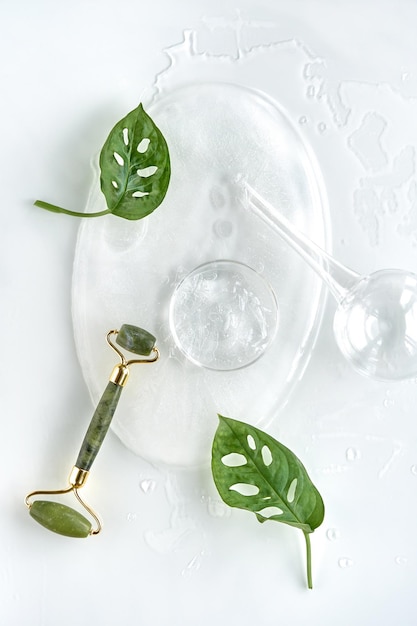 Moisturizer, green jade face roller on ice with ice cubes and exotic leaves. Monstera Adansonii leaves. Minimal flat lay on off white background. Facial massage with self made cosmetics.