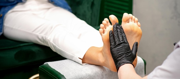 足に潤いを与えます。黒い手袋をしたペディキュアマスターの手は女性の足を気にします。足のマッサージ。ペディキュアビューティーサロンのコンセプト。
