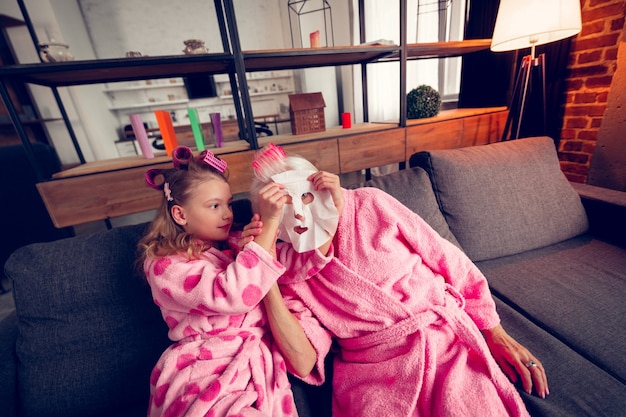 Moisturizing mask. Caring cute girl feeling involved while putting moisturizing mask on face of granny