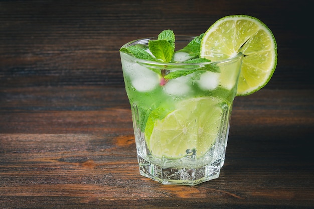 Mojito with mint and lime in a glass and a jug on the round board. Brown wood background.