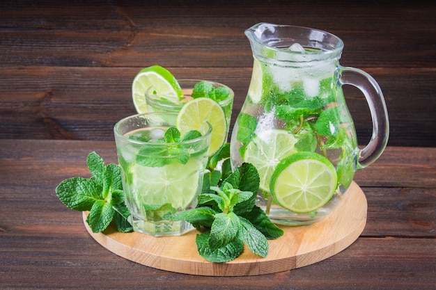 Mojito with mint and lime in a glass and a jug on the round board. Brown wood background.