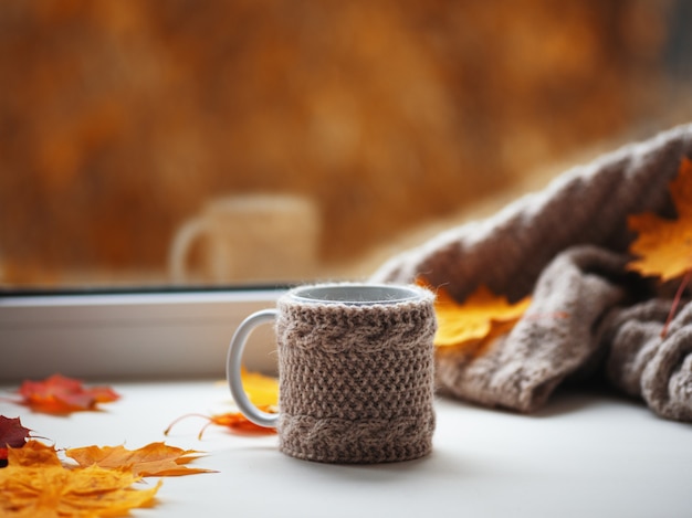 Mok warme koffie met gebreide warme trui en herfstbladeren op het raam in het huis. Hygge concept