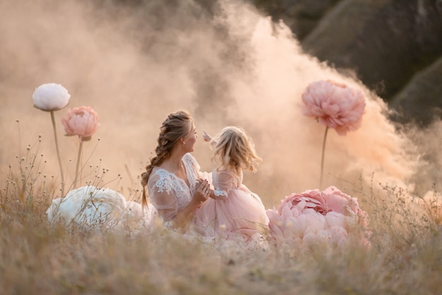 Фото Мама и дочка в розовых сказочных платьях смотрят вдаль, сидя в поле в окружении крупных розовых декоративных цветов