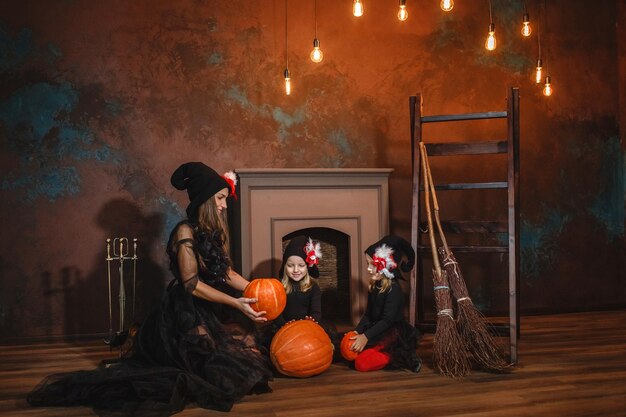 写真 魔女の衣装を着た母親と娘たちは ⁇ 暖炉の近くに座ってハロウィーンに南瓜で遊んでいます ⁇