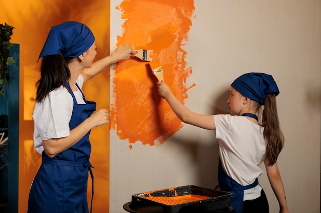 Мама и ребенок используют оранжевую краску на стенах и кисть, чтобы отремонтировать интерьер дома, вместе красят комнату в квартире. люди развлекаются ремонтом по дому, украшают своими руками.