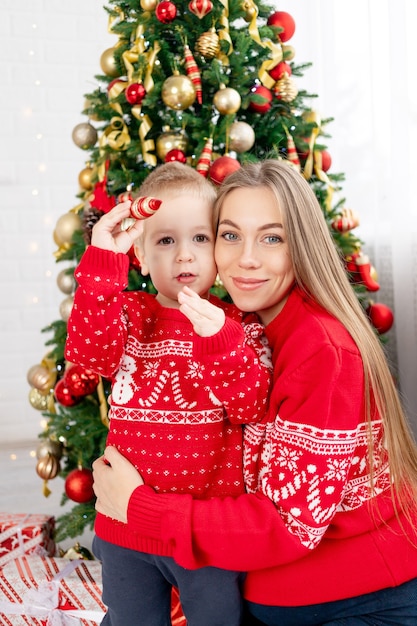 Mom and baby son in a red sweater under the Christmas tree at home are enjoying the new year and Christmas hugging and congratulating each other