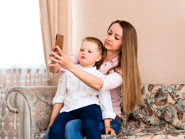 居心地の良いリビングルームで自分撮りをしているママと赤ちゃん。ママと息子は変な顔をして、カメラで自分の写真を撮ります