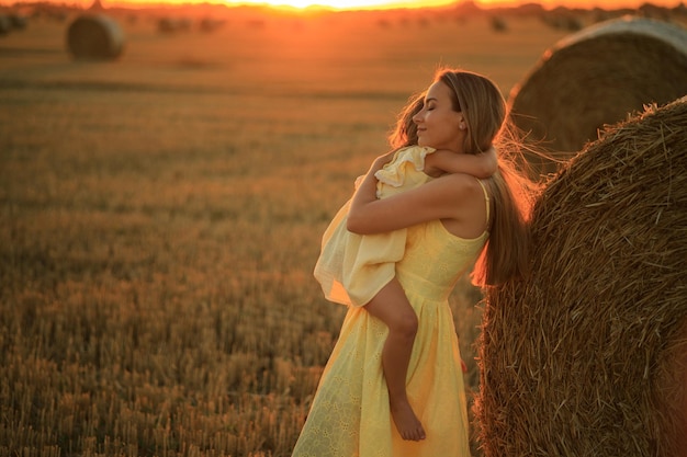 Mom and blonde daughter are sitting on a haystack in a field at sunset Mother and child have fun on the farm