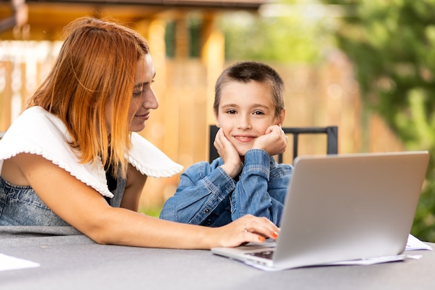 Mom and boy schoolboy are engaged in lessons through a laptop\
at home in the garden. online classes for children. schoolboy\
listens to a lecture and solves problems