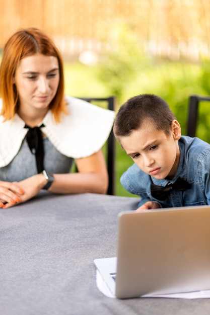 Mom and boy schoolboy are engaged in lessons through a laptop at home in the garden. Online classes for children. Schoolboy listens to a lecture and solves problems