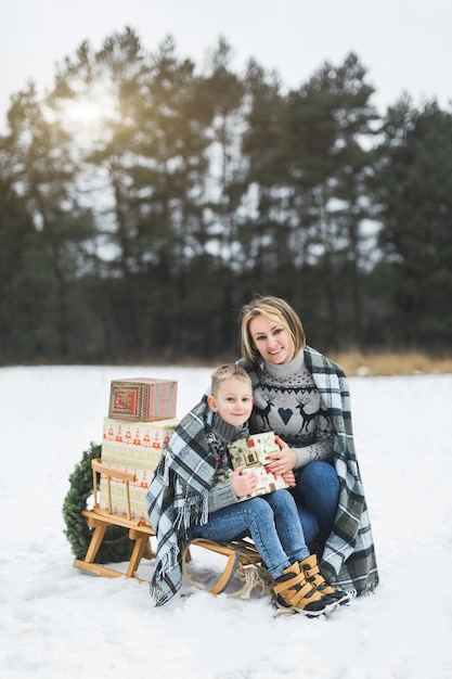 Mom and child sitting together on wooden decorated sledge with Christmas presents