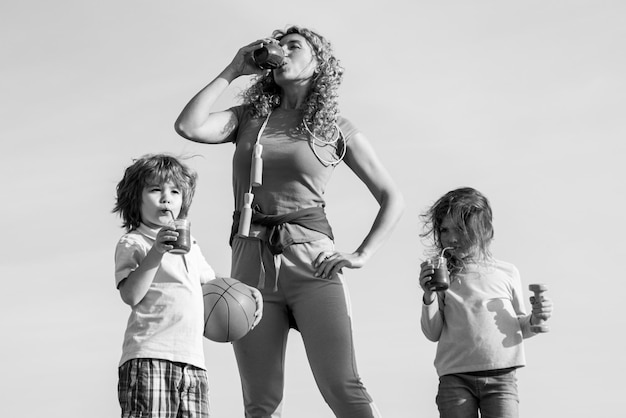 Mom and children drinking smoothie or juice Family after sport exercising on blue sky background copy space Happy laughing children and her beautiful young mother sporty loving family