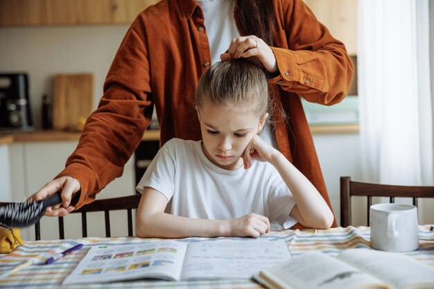 Mom combing her daughters hair while she does her homework at home at the kitchen table